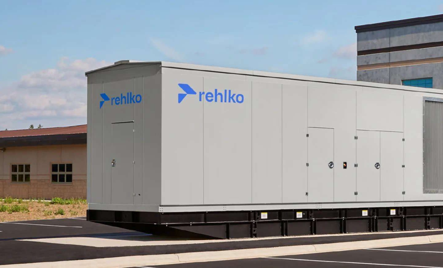 Large gray outdoor energy-system enclosure labeled “rehlko,” positioned next to a brick building under a clear blue sky.