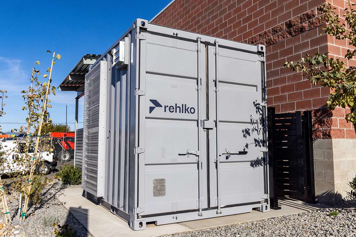 Large gray outdoor energy-system enclosure labeled “rehlko,” positioned next to a brick building under a clear blue sky.